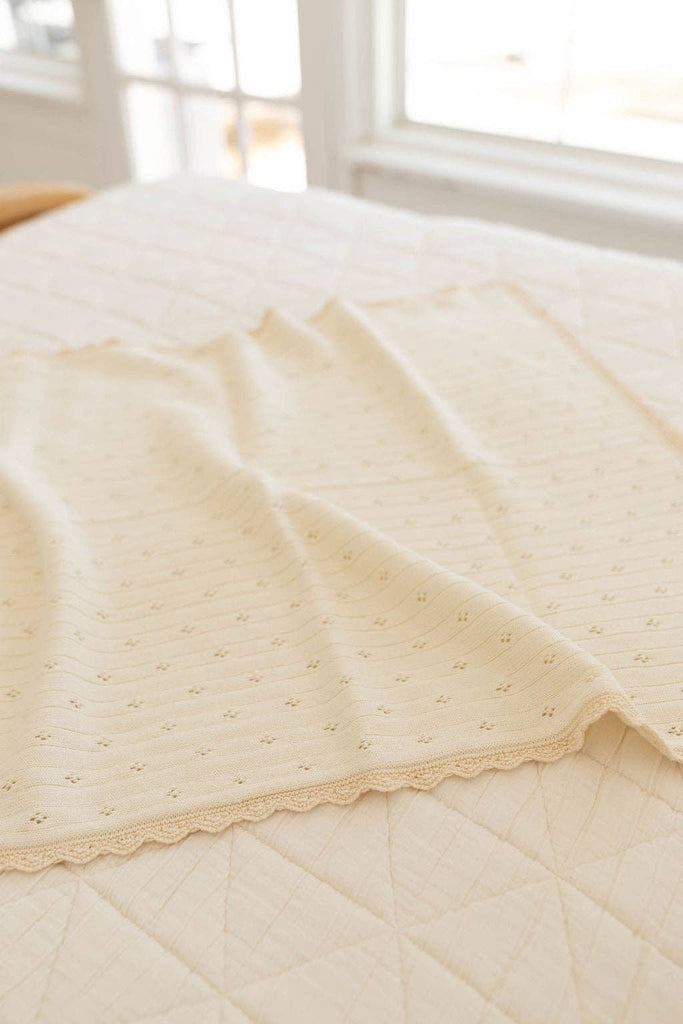 The Organic Pointelle Cotton Knit Baby Blanket, in a soft cream hue with a subtle dotted pattern and scalloped edges, is draped neatly over a quilted bed in a bright, sunlit room.