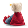 The 12-inch Steiff Superman Teddy Bear, with cream-colored fur, a bright red cape, red boots, a blue outfit, and a yellow tag on its left ear, is pictured sitting facing away from the camera.