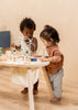 Two toddlers, one with curly hair and another with straight hair, are playing with a Wooden Activity Table at a small activity table in a warmly lit room.