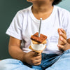 A child in a white t-shirt and jeans holds the Wooden Camp Fire Set s'more on a stick, complete with chocolate, marshmallow, and graham cracker pieces—ideal for indoor camping or kids' pretend play. The child's face is partially out of frame.