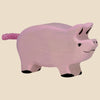 The Holztiger Piglet is a hand-carved pink wooden piglet with a curly tail, black hooves, and a small smile, shown on a beige background. Handcrafted in Europe and ships in one week.