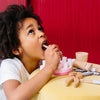 A young child plays with a Wooden Dentist Set, holding a wooden tongue depressor in their mouth at a yellow table with a red background.