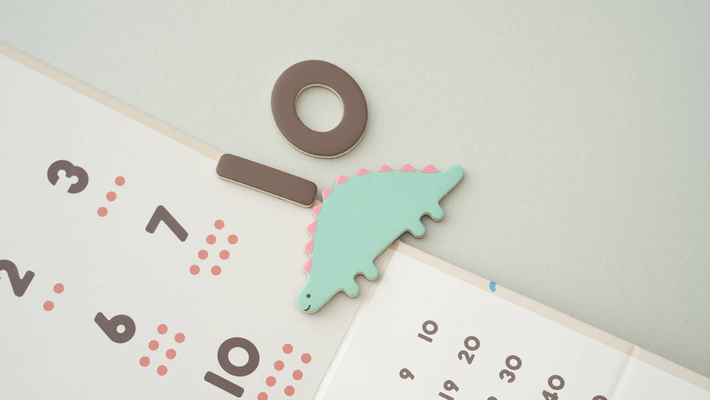 A pastel calendar with a green dinosaur magnet, brown circle and rectangle magnets clipped on top—perfect for pairing with the Magnetic Number Play Set or fun magnet puzzles.