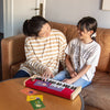 A woman and a child sit on a brown couch, smiling at each other. The child plays a small red Loog Piano with velocity-sensitive keys on a table covered with colorful cards.