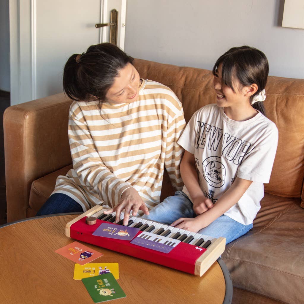 A woman and a child sit on a brown couch, smiling at each other. The child plays a small red Loog Piano with velocity-sensitive keys on a table covered with colorful cards.
