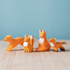 Four orange wooden fox figures in various sizes and poses are displayed on a light wooden surface with a blue background—perfect as the Bumbu Handmade Wooden Fox Family Set of charming handmade toys.