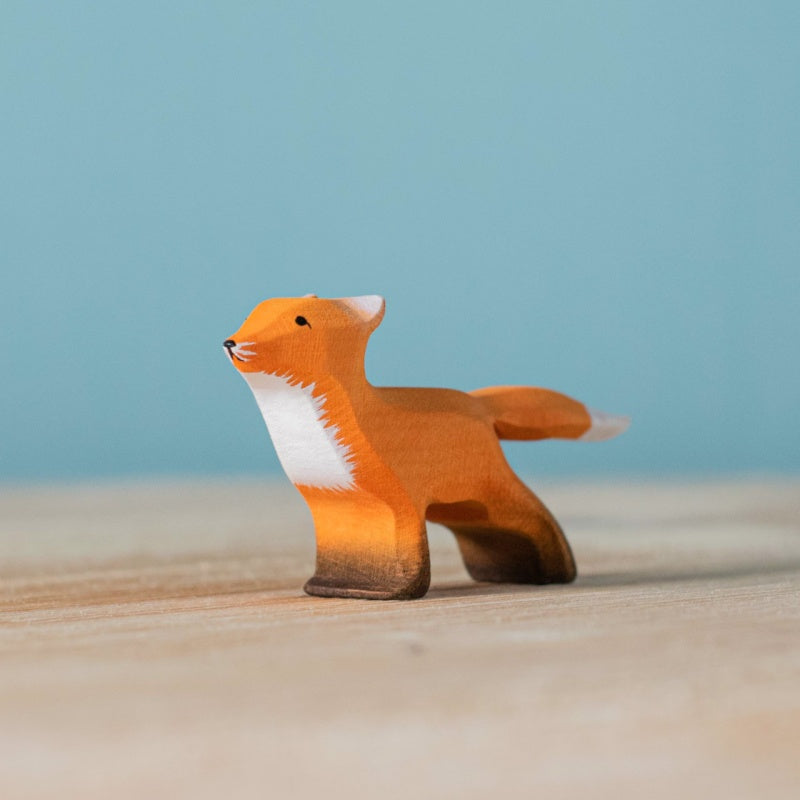 The Bumbu Handmade Wooden Fox Family Set includes a small wooden fox figurine, handcrafted in Romania with orange and white painted details, displayed on a light wood surface against a blue background.