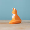 A small figurine from the Bumbu Handmade Wooden Fox Family Set, featuring orange and white details, sits on a light wooden surface against a plain light blue background.