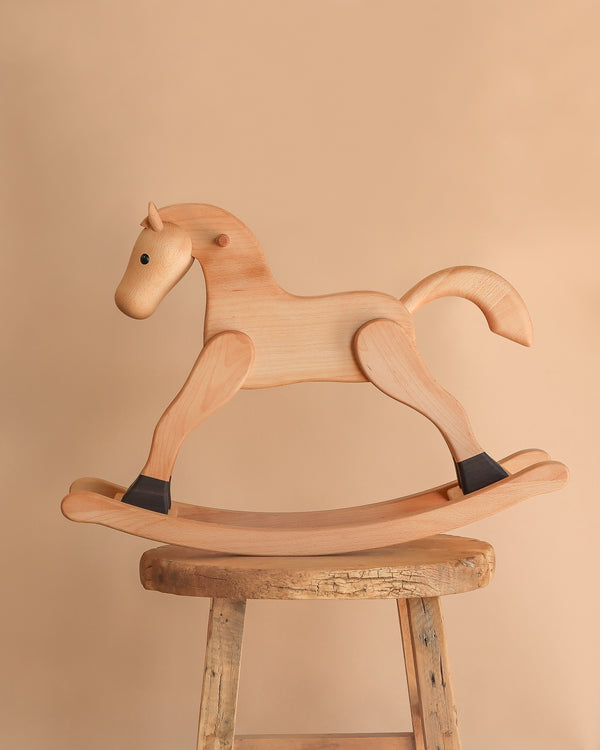 The Spring Copenhagen Wooden Rocking Horse – Final Sale features black hoof accents and minimalist Nordic design, elegantly displayed atop a rustic stool with a beige backdrop.