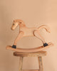 The Spring Copenhagen Wooden Rocking Horse – Final Sale features black hoof accents and minimalist Nordic design, elegantly displayed atop a rustic stool with a beige backdrop.
