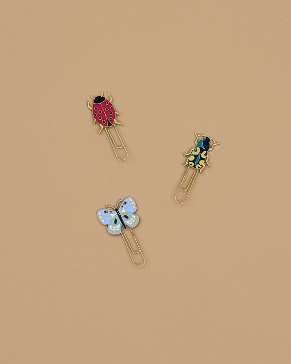 The Curio Enamel Paper Clip Set features three clips—a red ladybug, yellow and black beetle, and light blue butterfly—arranged on a beige background. Each measures about 4 x 4 x 0.5 in (10.2 x 10.2 x 1.3 cm).