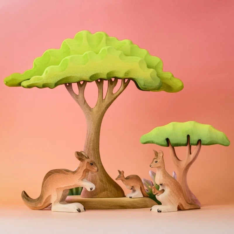 The Bumbu Wooden Kangaroo Family Set, crafted from sustainably sourced wood, is arranged with two stylized trees on a pink and orange gradient background for a playful, natural look with soft colors and smooth shapes.