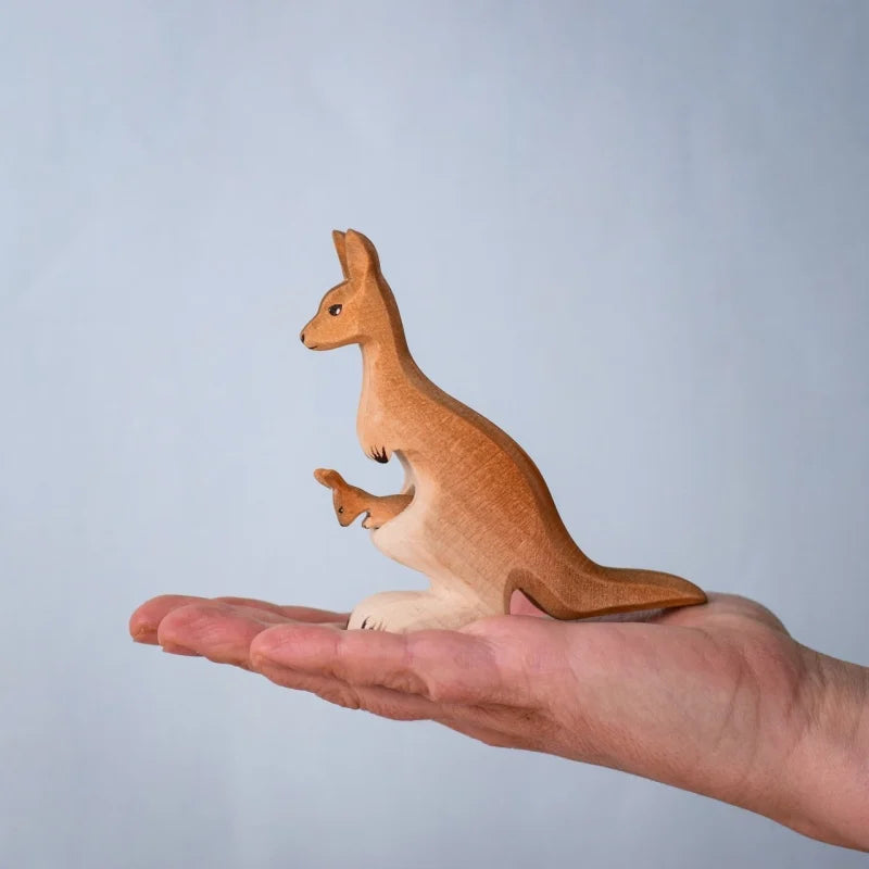 A hand holds the Bumbu Wooden Kangaroo Family Set, a small figurine made from sustainably sourced wood, featuring a kangaroo with a joey in its pouch against a plain light blue background.