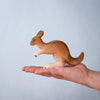 A hand holds a small brown and white kangaroo figurine from the Bumbu Wooden Kangaroo Family Set, made from sustainably sourced wood, set against a plain light blue background.