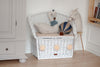 A white wicker bench with a cushion, decorated with a swan plush, llama doll, and the Hot Air Balloon - Light Brown / Navy Blue, sits in the corner of a children's room with light wood floors and neutral furniture.