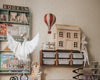 A cozy children's room featuring shelves of books, a wooden dollhouse, storage bins, a hanging plush swan, the Hot Air Balloon - Burgundy decor, a tiger-shaped cutout, and a doll with a pointed hat sitting on a shelf.