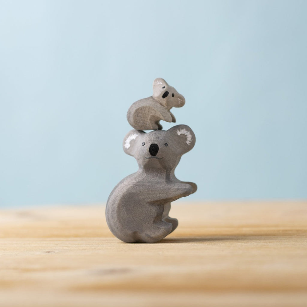 The Bumbu Handmade Wooden Koala Bears with Eucalyptus Tree Set features a small wooden koala and baby on a light surface with a blue background, perfect for Montessori play or as an addition to a wooden animal collection.