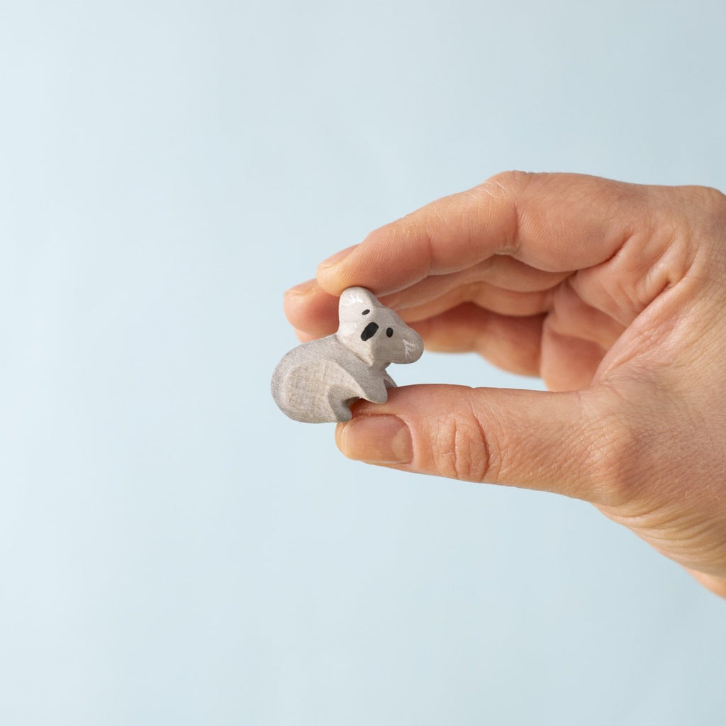 A hand holds a Bumbu Handmade Wooden Koala Bears with Eucalyptus Tree Set figurine against a plain light blue background, ideal for Montessori toys or enhancing your wooden animal collection.