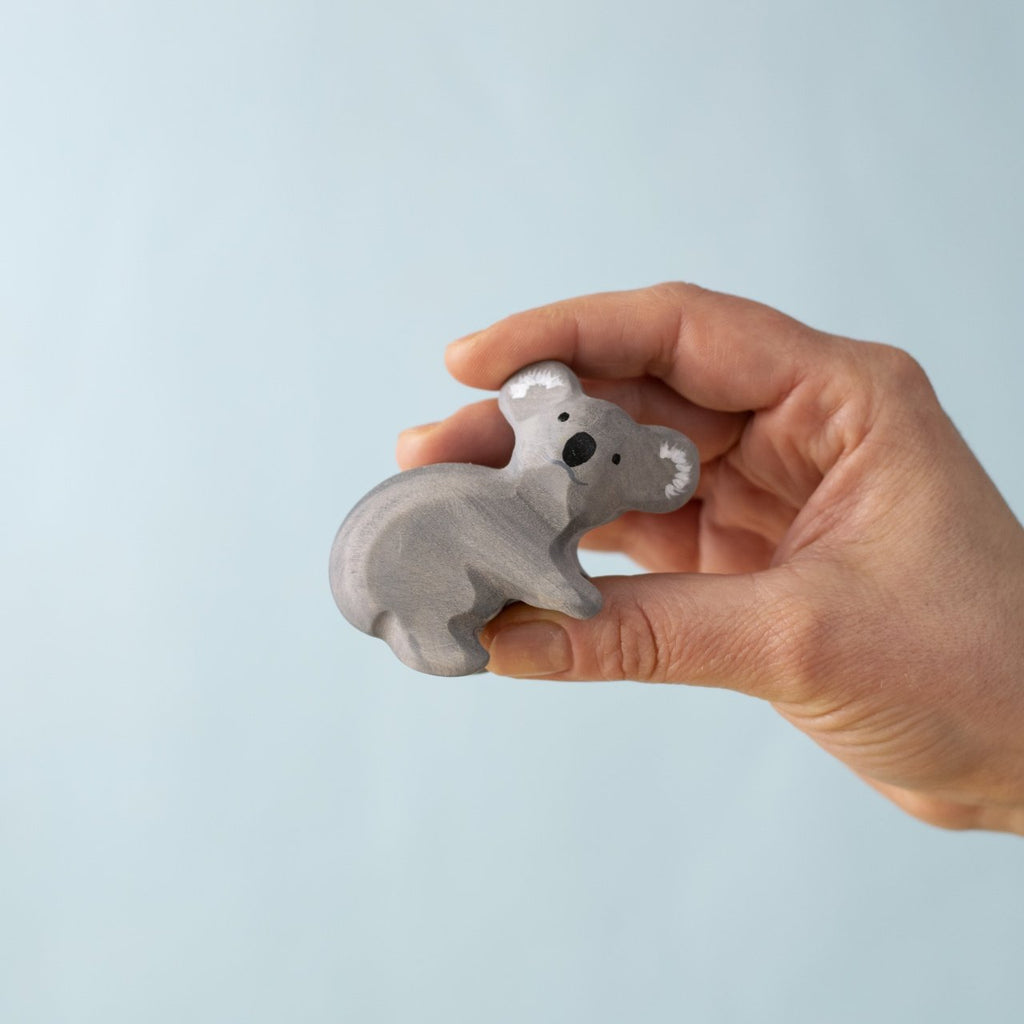 A hand holds a small, gray Bumbu Handmade Wooden Koala Bear from the Koala Bears with Eucalyptus Tree Set, shown against a light blue background—ideal for Montessori play or koala collections.