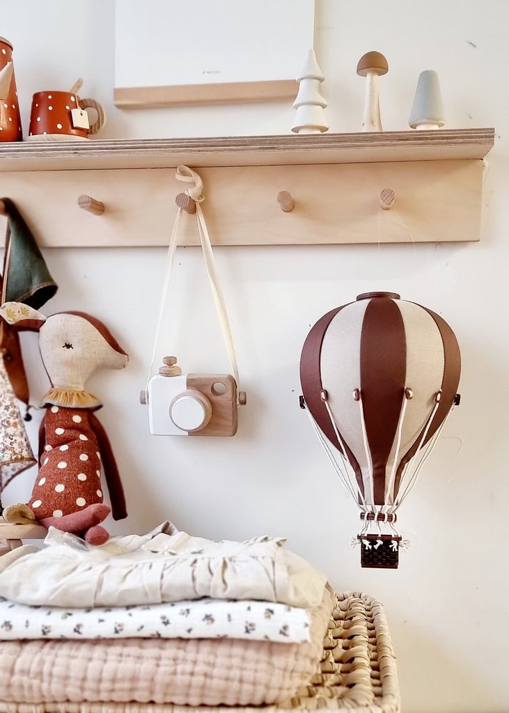 A cozy nursery scene with a plush duck toy, wooden toy camera, folded baby clothes, and a Hot Air Balloon - Light Brown / Dark Brown wall decor, plus a shelf holding wooden ornaments and polka-dot mugs on a beige wall.