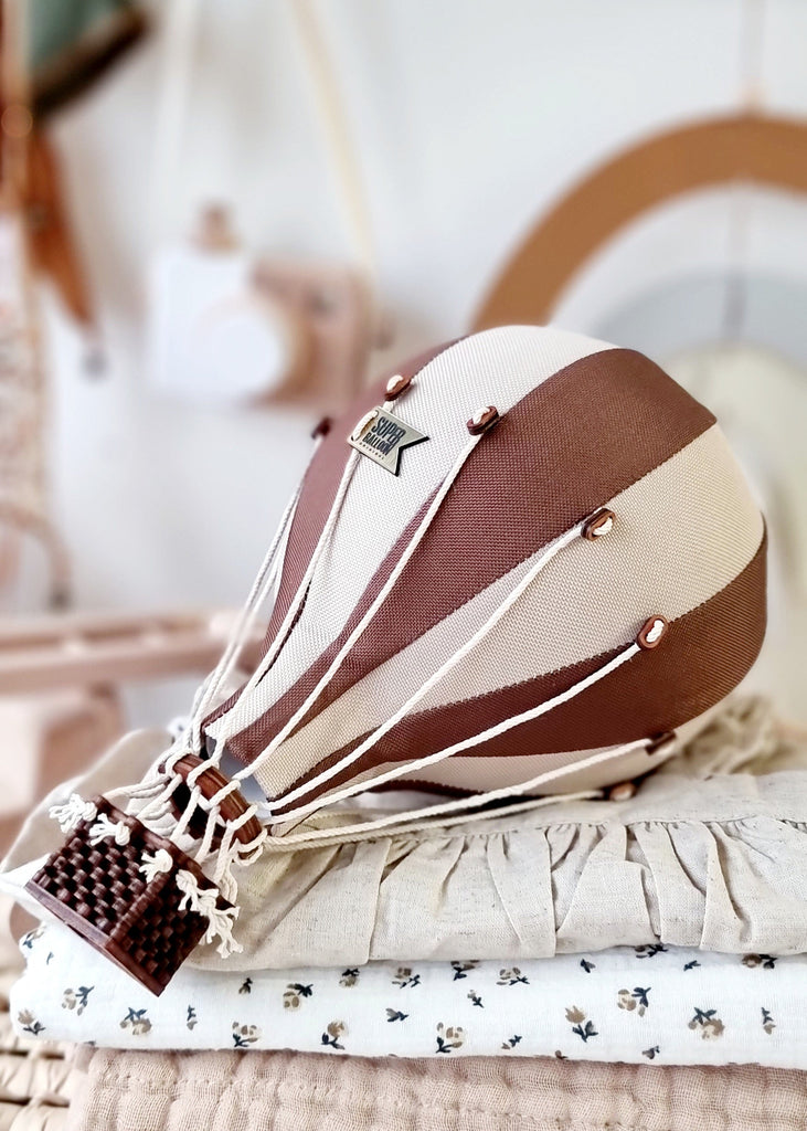 The Hot Air Balloon - Light Brown / Dark Brown model features brown and beige stripes and sits on textured fabric with a soft background—an ideal, charming decoration for any child's room.