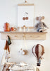 A wooden shelf displays a red teapot set, white cone trees, a cherry print, and a stuffed mouse. Below hang a toy camera, scarf, small wooden airplane, and Hot Air Balloon - Light Brown / Dark Brown for children's room decor.