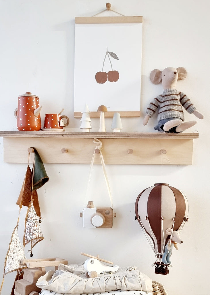 A wooden shelf displays a red teapot set, white cone trees, a cherry print, and a stuffed mouse. Below hang a toy camera, scarf, small wooden airplane, and Hot Air Balloon - Light Brown / Dark Brown for children's room decor.