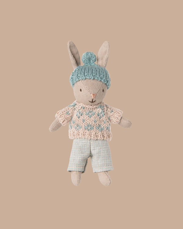 PRE-ORDER: Winter Rabbit, Micro - Sand - Mint features a soft rabbit toy in a blue hat with pom-pom, pink and blue sweater, and blue plaid pants—perfect for any miniature dollhouse. Estimated Ship Date: Early November. Set against a beige background.