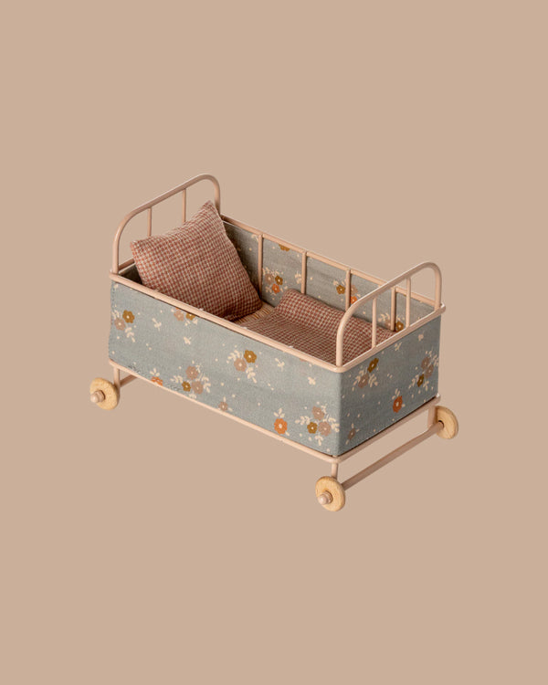 The PRE-ORDER: Cot Bed, Micro - Powder features a pink frame with floral fabric and includes two checkered pillows. Shown against a plain beige background. Estimated ship date: Early October.