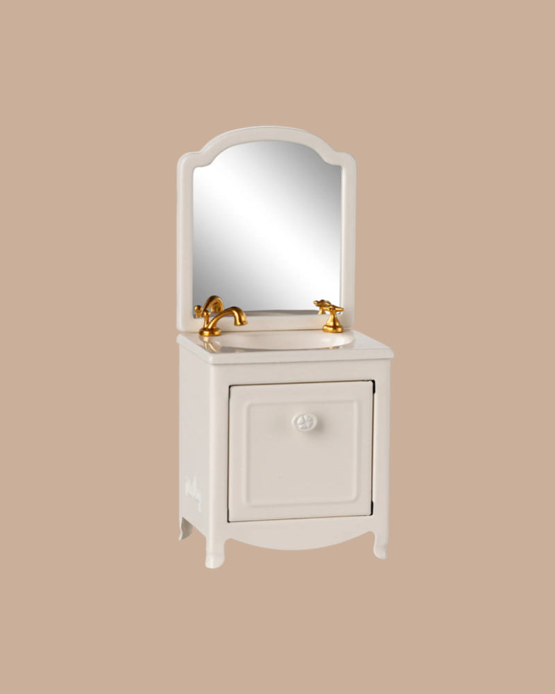The PRE-ORDER: Sink Dresser, Mouse - Off White (ships early September) features a small white vanity with a cabinet, gold faucets, and an arched mirror styled with bathroom essentials against a beige background.