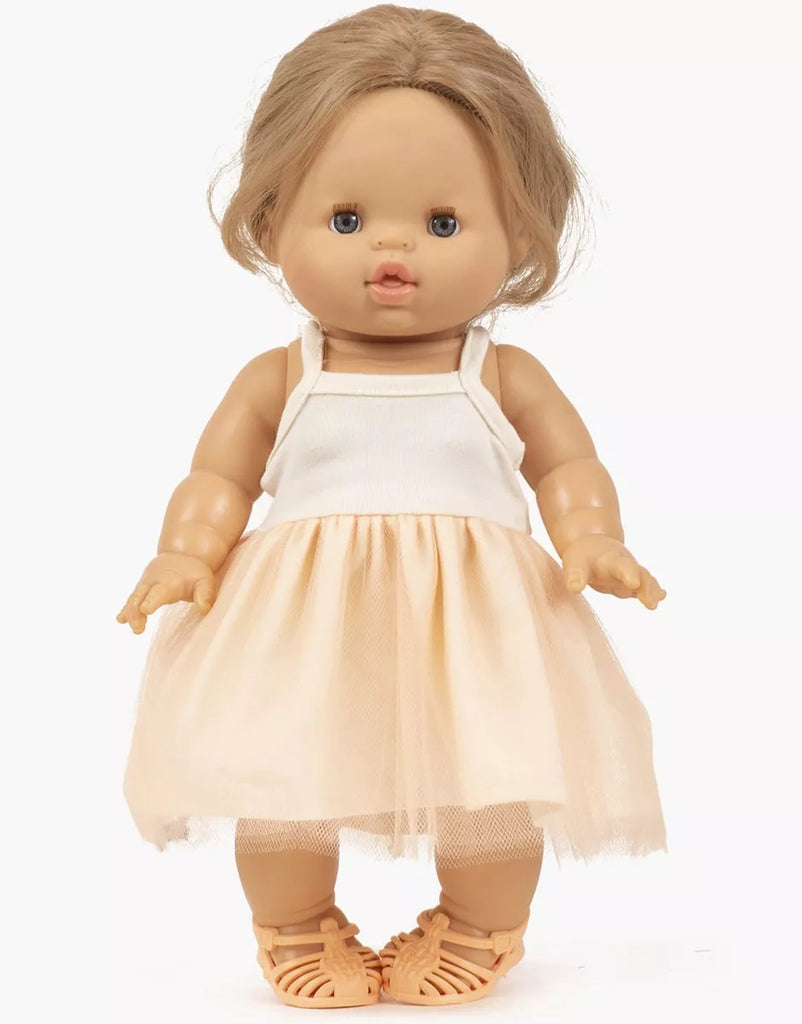 A Minikane doll with light brown skin, long blonde hair, and blue eyes wears a white tank top, peach tulle skirt, and Minikane Beach Sandals - Peach, standing against a plain white background.