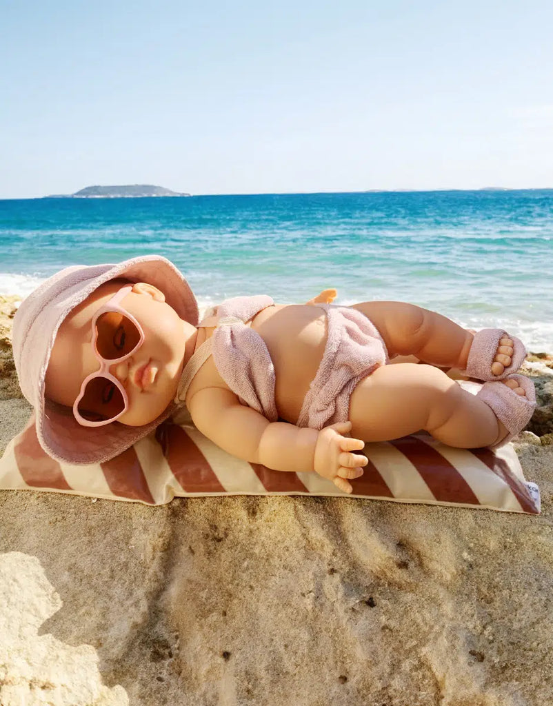 A Minikane doll wears the Emile & Ida Ursula 3-piece Terrycloth Swimsuit in Petal, relaxing on a striped pillow at the beach with blue waves and an island in the background.