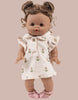 A Minikane doll wears the Emile & Ida Emilia Dress in Soft Terrycloth - Flower, a cream outfit with small prints, and pink sandals. The brown-skinned doll features curly hair in two buns and stands against a beige background. Designed in France.
