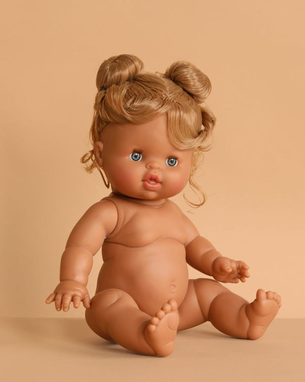 The Minikane Baby Doll (13") - Louise features light brown skin, blue eyes, and curly blonde hair in two buns. This anatomically correct doll sits upright against a beige background. Clothing sold separately—perfect for realistic play.