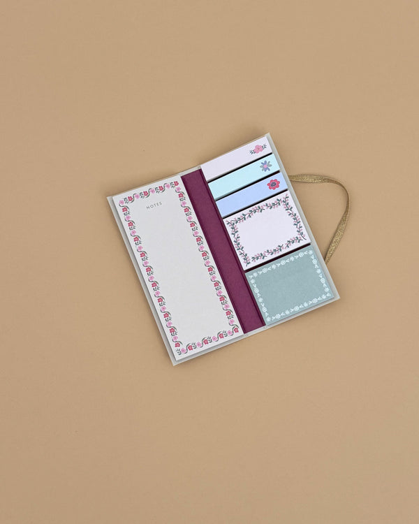 The Delphine Sticky Note Folio, featuring floral-patterned sticky notes and memo pads in assorted sizes and adorned with a gold ribbon, is displayed open on a plain light brown surface.