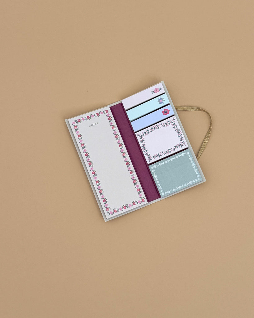 The Delphine Sticky Note Folio, featuring floral-patterned sticky notes and memo pads in assorted sizes and adorned with a gold ribbon, is displayed open on a plain light brown surface.