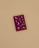 The Gracie Mini Embroidered Journal, a red hardcover with floral and berry embroidery, rests on a plain light brown surface and features blank unlined pages for creative expression.