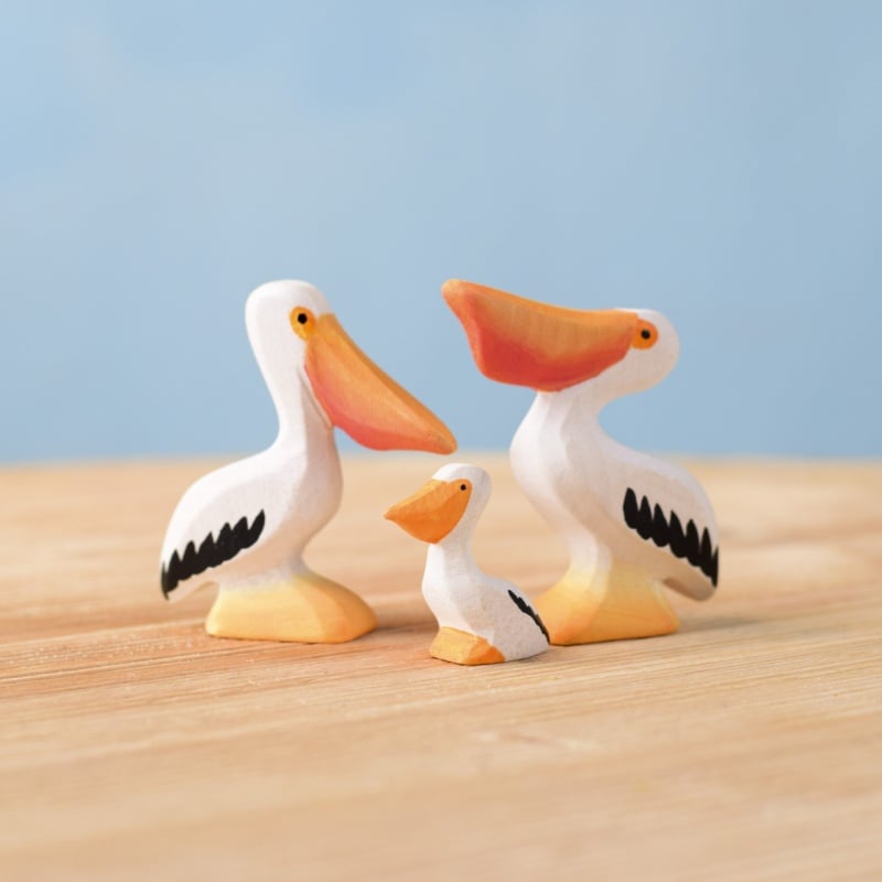The Bumbu Handmade Wooden Pelicans Family Set features three white wooden pelican toys with black and orange details, arranged on a light wooden surface against a blue background—ideal for Montessori play and sustainable toy collections.