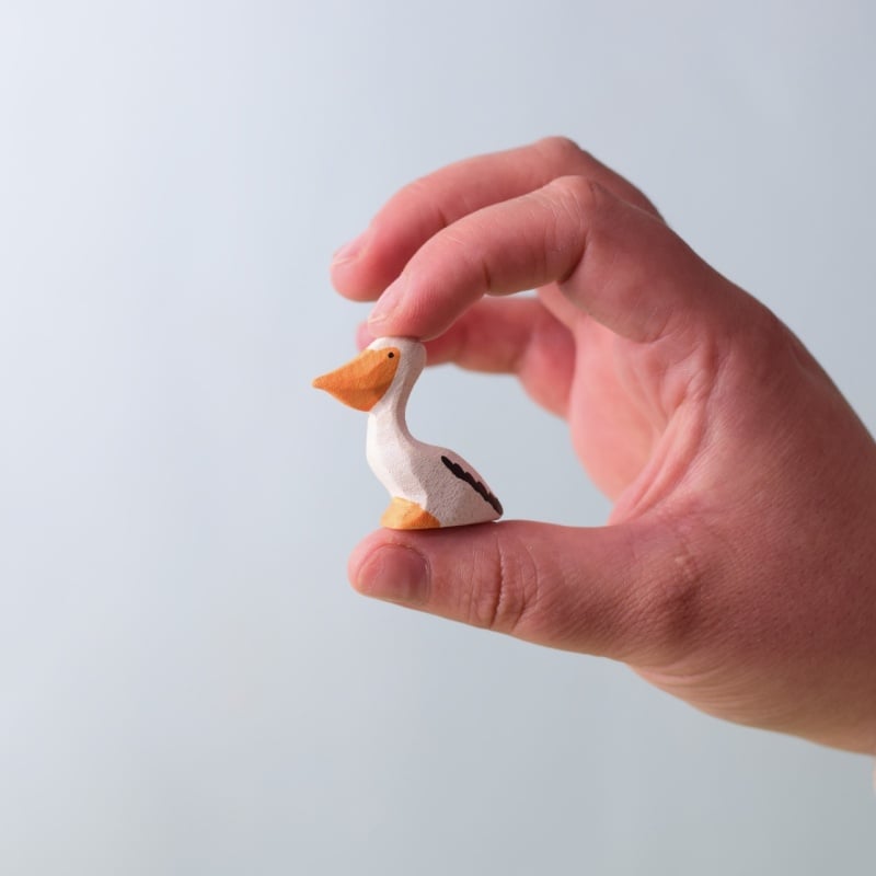 A hand delicately holds a Bumbu Handmade Wooden Pelicans Family Set toy, featuring an orange beak and white body—perfect for Montessori play—against a softly blurred, pale background.