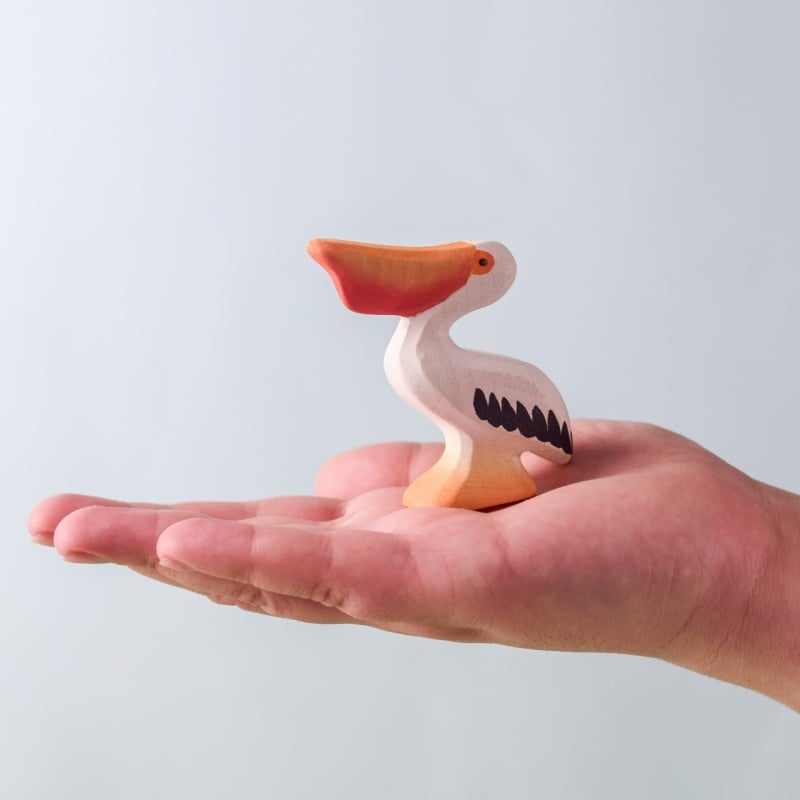 A hand holds a small, colorful pelican from the Bumbu Handmade Wooden Pelicans Family Set, featuring an orange beak, white body, and black wing accents—an adorable sustainable wood toy—against a plain light background.