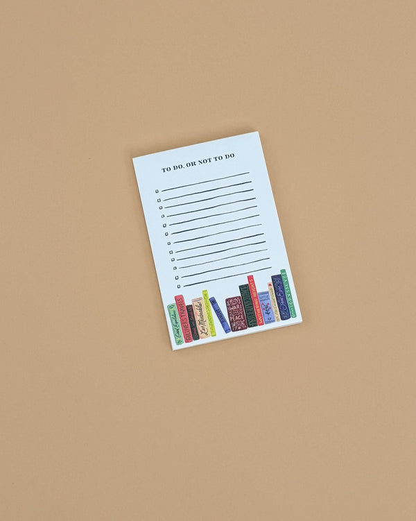 The "To Do, Or Not To Do Notepad" features lined spaces for your tasks and colorful illustrated books at the bottom, all on a beige background. This charming tear-off notepad is perfect for organizing your to-do list with style.