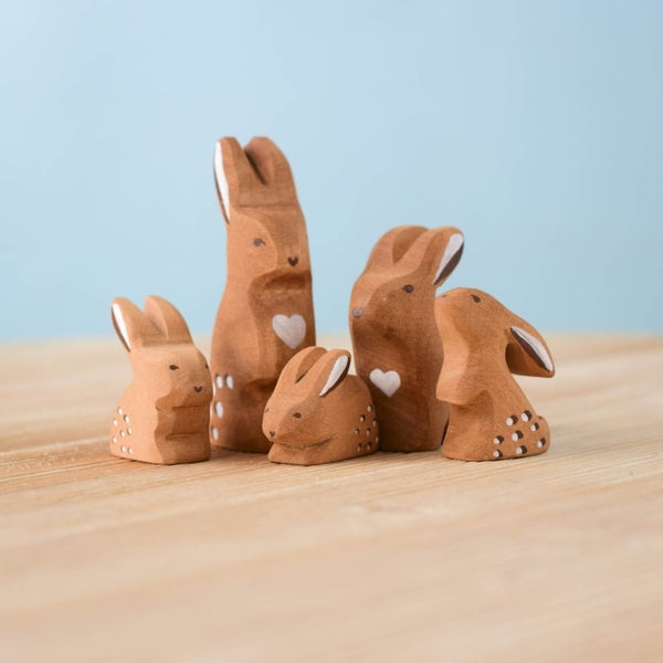 The Bumbu Handmade Wooden Brown Bunny Rabbit Family Set features six small, handcrafted wooden bunny figurines with white details and heart shapes, arranged on a light wood surface against a pale blue background—perfect for Montessori collections.