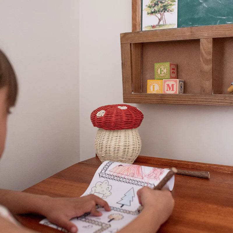 A child colors at a wooden desk with the Olli Ella Mushroom Basket With Coloring Pencils- Red. Twig pencils and the woven basket are on the desk, with wooden alphabet blocks and a small shelf above.