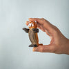 A hand holds a small, handcrafted Bumbu Wooden Red Panda from the Red Panda Family with Bamboo Set, featuring painted eyes and facial details, set against a light blue background.