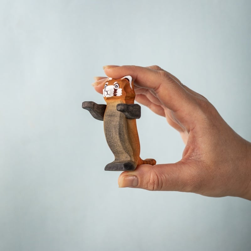 A hand holds a small, handcrafted Bumbu Wooden Red Panda from the Red Panda Family with Bamboo Set, featuring painted eyes and facial details, set against a light blue background.
