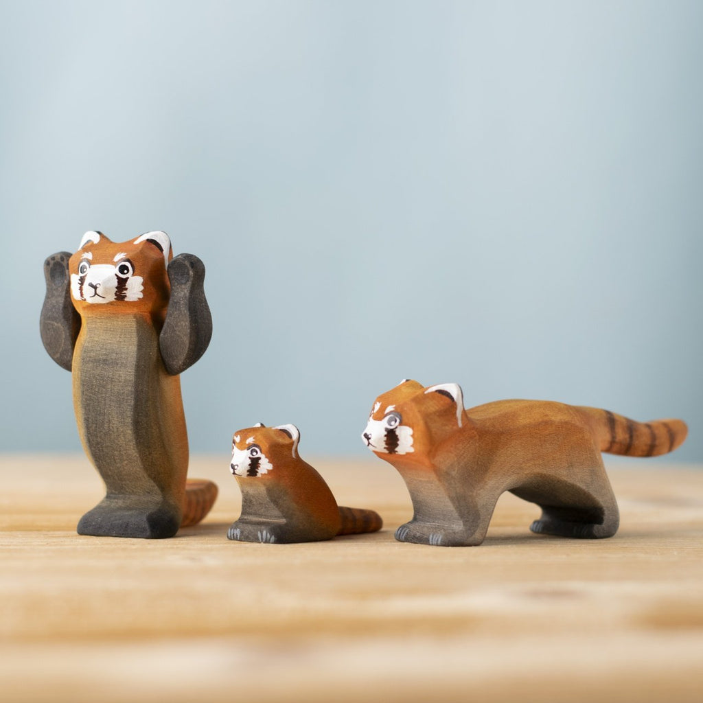 Three Bumbu Handmade Wooden Red Panda Family with Bamboo Set figures are displayed on a wooden surface—a standing adult with raised arms and two smaller red pandas on all fours—set against a plain, light blue background.