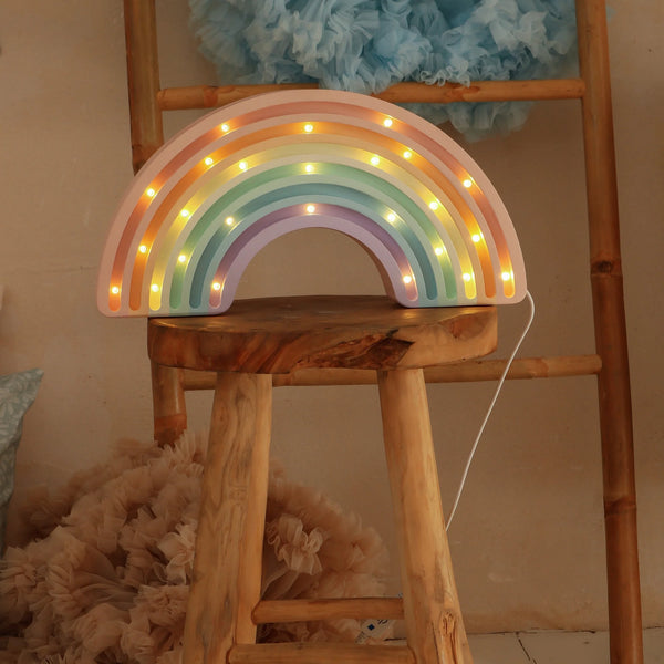 The Little Lights Rainbow Lamp, a handmade children's night light with glowing pastel colors, sits on a wooden stool in front of a ladder adorned with fluffy blue and beige fabric.