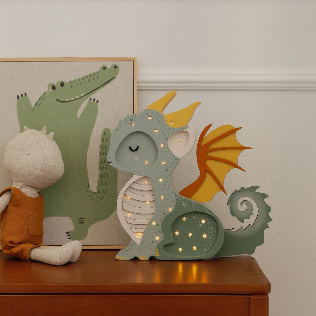 A Handmade Wooden Lamp - Dragon, Hunter Green sits on a dresser, adding whimsical nursery decor beside a dragon toy and a framed illustration of a smiling green crocodile.