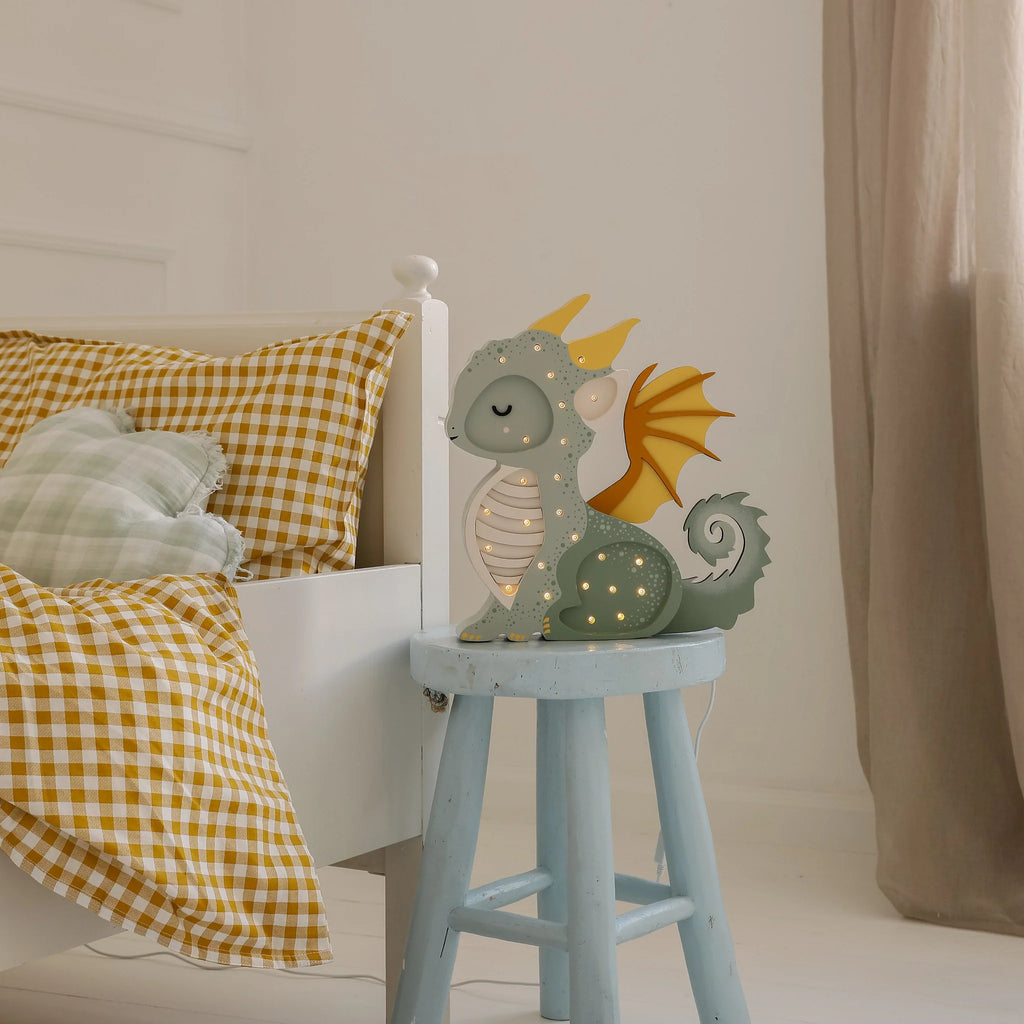 The Handmade Wooden Lamp - Dragon, Hunter Green sits on a blue stool by a bed with yellow gingham bedding and plaid pillows, making charming nursery decor and a delightful children's night light in this cozy bedroom.