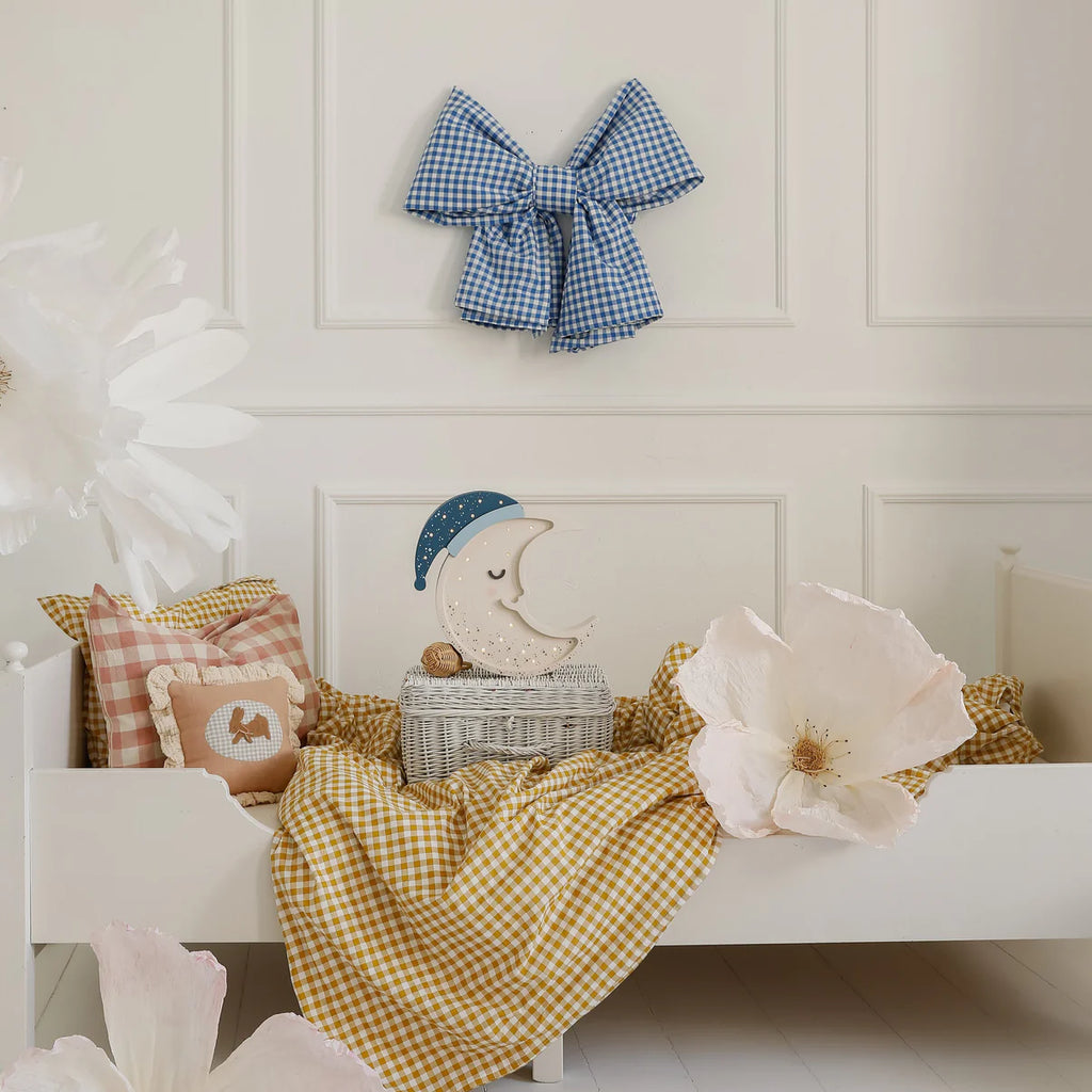 A cozy child's bed with a yellow gingham blanket, pink checkered pillows, large white paper flowers, and a Handmade Wooden Lamp - Moon as a night light on a wicker basket—a perfect nursery decor for sweet dreams.